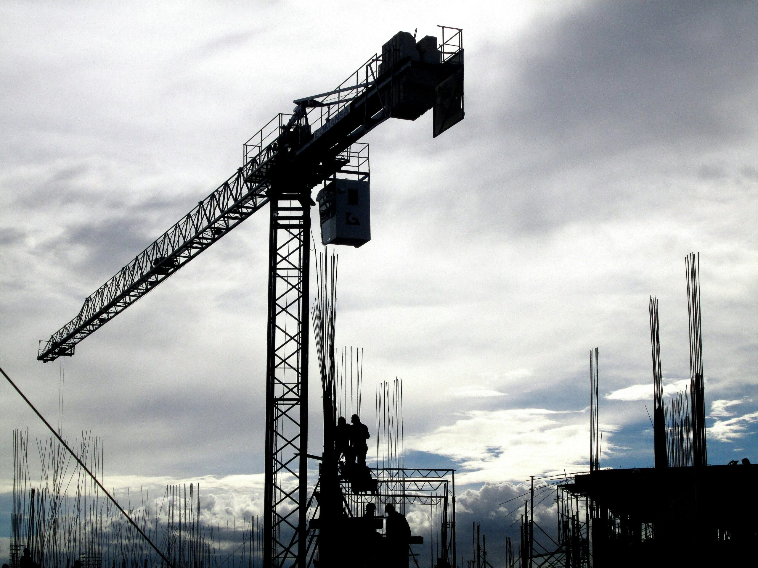 Silhouetted construction crane and workers against a cloudy sky during the day.